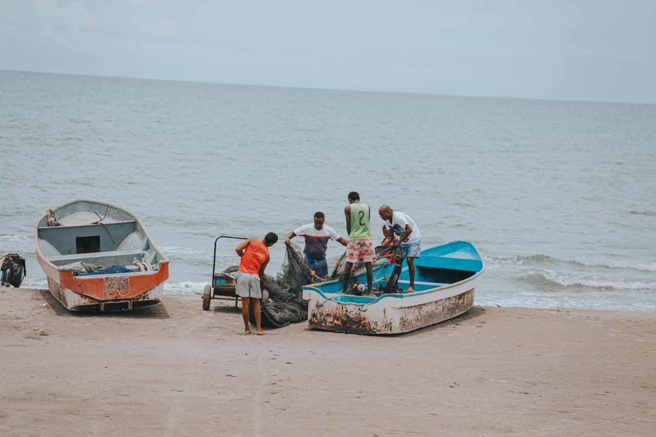Aparecen tres lanchas a la orilla del mar, sobre dos de ellas cinco hombres afrodescendientes desenredando una mantarraya y de fondo el mar del océano pacífico.
