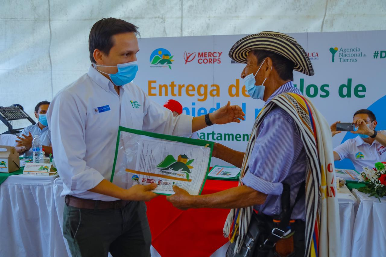Funcionario de la ANT, vestido de camisa blanca, pantalón azul y tapabocas, hace entrega título de propiedad a un campesino, quien lleva un sombrero, poncho, carriel y tapabocas. De fondo en mesa cubierta con mantel blanco otros funcionarios  de la ANT  y una pancarta de la agencia. 
