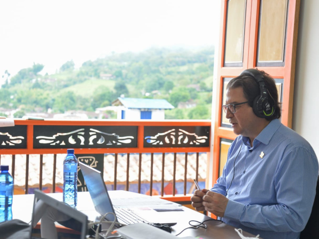 El Ministro de Agricultura y Desarrollo Rural, Rodolfo Zea Navarro, aparece sentado, frente tiene un portátil, lleva puestos unos audífonos grandes. Sobre la mesa dos botellas de agua, detrás, una ventada en madera abierta y de fondo una montaña y una casa.