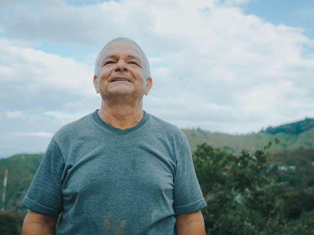 Aparece un señor, adulto mayor, de medio cuerpo, al aire libre, sonriente, con una camiseta color azul y de fondo las montañas verdes.