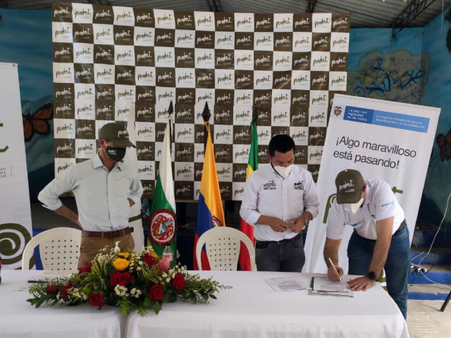 Tres hombres del equipo de la ANT en mesa de conversación, cada uno con tapabocas y camisa blanca, uno de ellos firmando un documento sobre la mesa. Encima de la mesa un arreglo de flores.