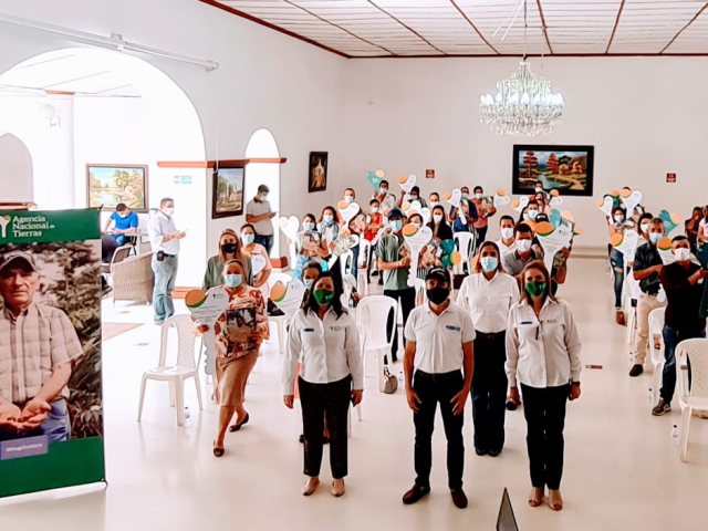 Varios campesinos víctimas de la violencia en la subregión del Catatumbo, están de pie, a su lado tienen una silla blanca. En la parte de adelante un pendón del la ANT en donde aparece un señor, campesino, de la tercera edad.