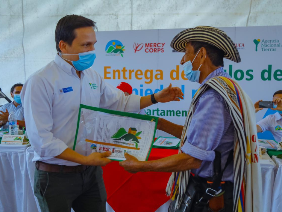 Funcionario de la ANT, vestido de camisa blanca, pantalón azul y tapabocas, hace entrega título de propiedad a un campesino, quien lleva un sombrero, poncho, carriel y tapabocas. De fondo en mesa cubierta con mantel blanco otros funcionarios  de la ANT  y una pancarta de la agencia. 
