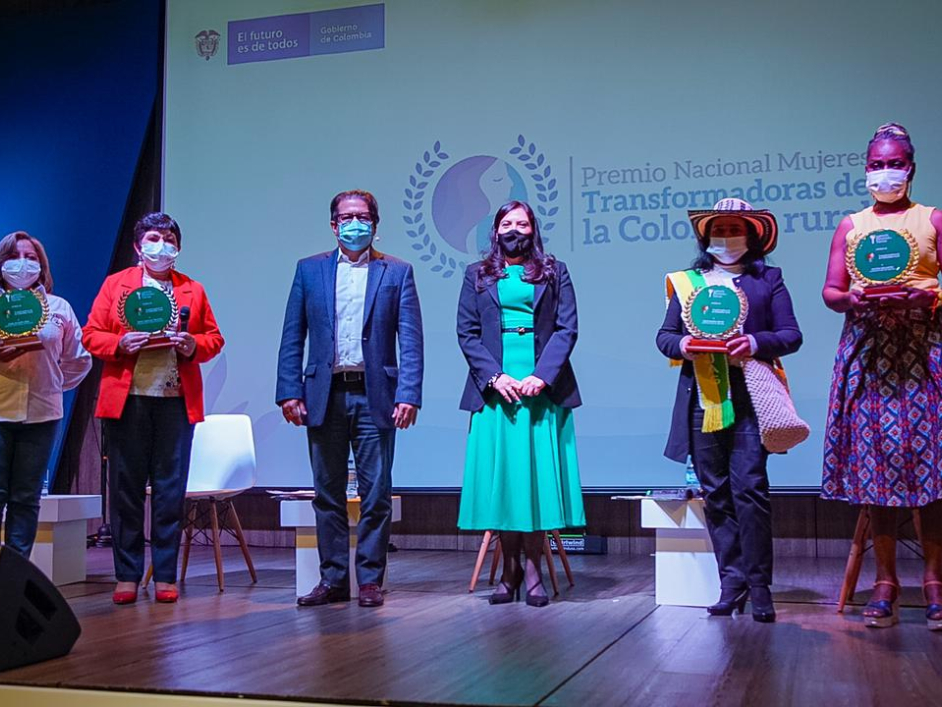 Fotografía de la celebración y entrega del premio nacional MUJERES TRANSFORMADORAS DE LA COLOMBIA RURAL. Sobre una tarima con piso de madera y de fondo el logo de la premiación, están cuatro mujeres ganadoras de este premio en compañía de la Directora de la ANT y un funcionario hombre de la ANT. 
