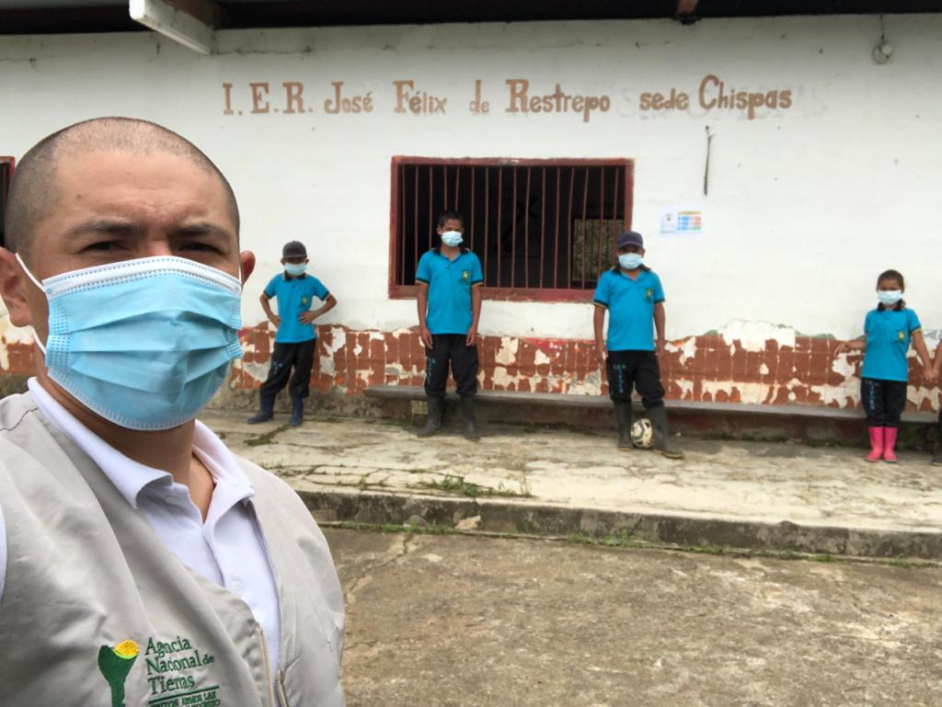 Selfi tomada por un hombre funcionario de la ANT. Él de medio cuerpo, luce el chaleco corporativo de la ANT y lleva puesto camisa blanca y tapabocas. De fondo aparece la institución educativa José Félix Restrepo sede Chispas y cuatro niños estudiantes vistiendo todos camisas iguales de color azul, pantalón negro y botas de caucho. 