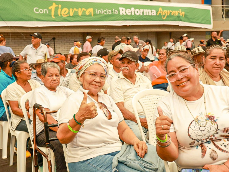 Mujeres asistentes al evento, sonriendo para la cámara, con un cartel detrás que dice "Con tierra inicia la reforma agraria"