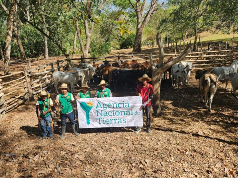 Campesinos de pie frente a una cerca con ganado y un cartel de la ANT.
